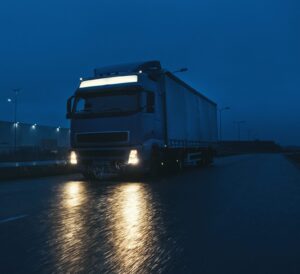 Blue Long Haul Semi-Truck with Cargo Trailer Full of Goods Travels At Night on the Freeway Road, Driving Across Continent Through Rain, Fog, Snow. Industrial Warehouses Area. Front Shot