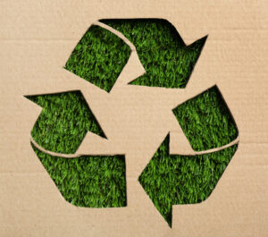 Sheet of cardboard with cutout recycling symbol on green grass, top view