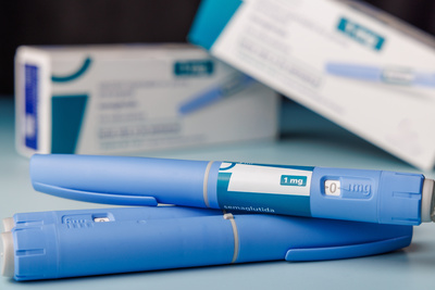 Two blue semaglutide injection pens lying on a light blue surface with blurred medication boxes in the background, labeled "1 mg semaglutida."
