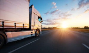A large white transport truck driving on a highway during a bright sunset, representing long-distance medication delivery.