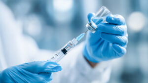 A close-up of hands in blue medical gloves using a needle to draw clear liquid medication from a glass vial into a syringe.