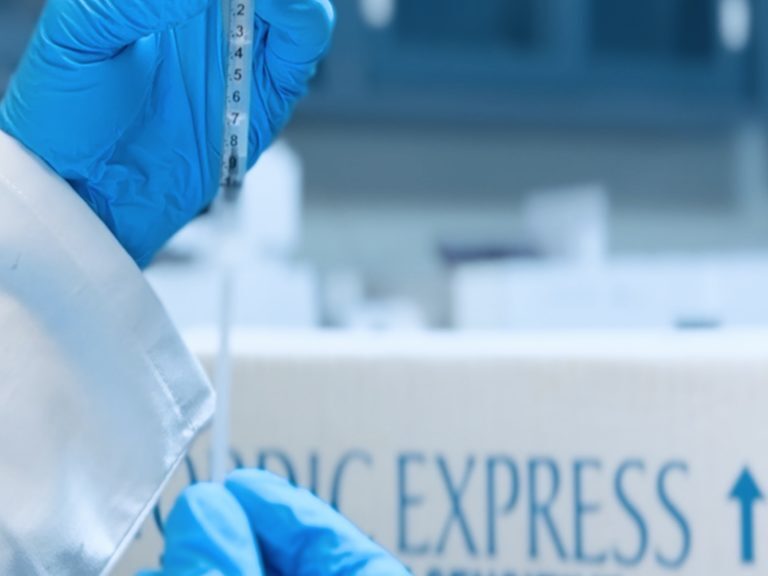 A side view of a medical professional in a lab coat and blue gloves handling a syringe in front of a Nordic Express shipping box.