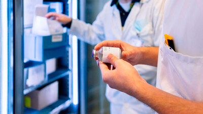 Close-up of a pharmacist's hands holding a medical vial in front of an open specialized medical refrigerator with blue LED lighting and organized medicine bins.