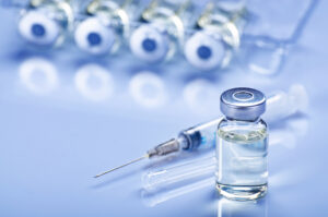 A single clear medication vial with a silver cap sits in sharp focus in the foreground, with a medical syringe and a blurred row of several other vials in the background under blue-toned lighting.