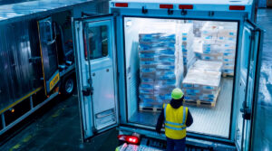 An aerial view inside a cool-toned warehouse where a delivery driver, wearing a yellow safety vest and helmet, stands at the back of an open refrigerated truck fully loaded with small-format medication boxes on pallets, emphasizing integrated distribution.