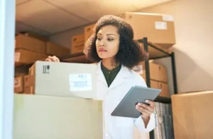 A professional female pharmacist with curly hair, wearing a white coat and blue gloves, carefully inspects a shipping box label while holding a tablet in a well-organized stockroom.
