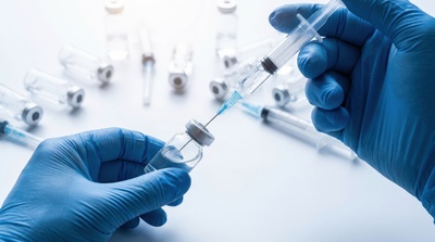 Close-up of hands in blue medical gloves carefully withdrawing liquid from a glass vial using a needle and syringe, with several other empty vials scattered on a white surface.
