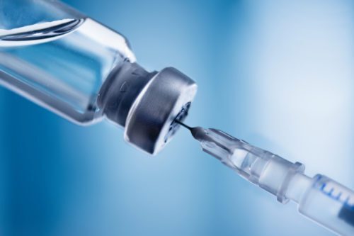 A dramatic close-up angle of a clear syringe needle piercing the rubber stopper of an inverted glass medicine vial against a soft blue background. Clear liquid is visible inside the vial.